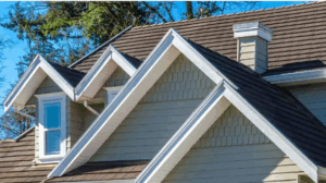 Close-up view of a house exterior with multiple peaked gable roofs, gray siding, white trim, and a small window under one gable.