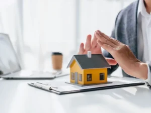 A person places protective hands over a small model house on top of documents, suggesting home security or insurance.